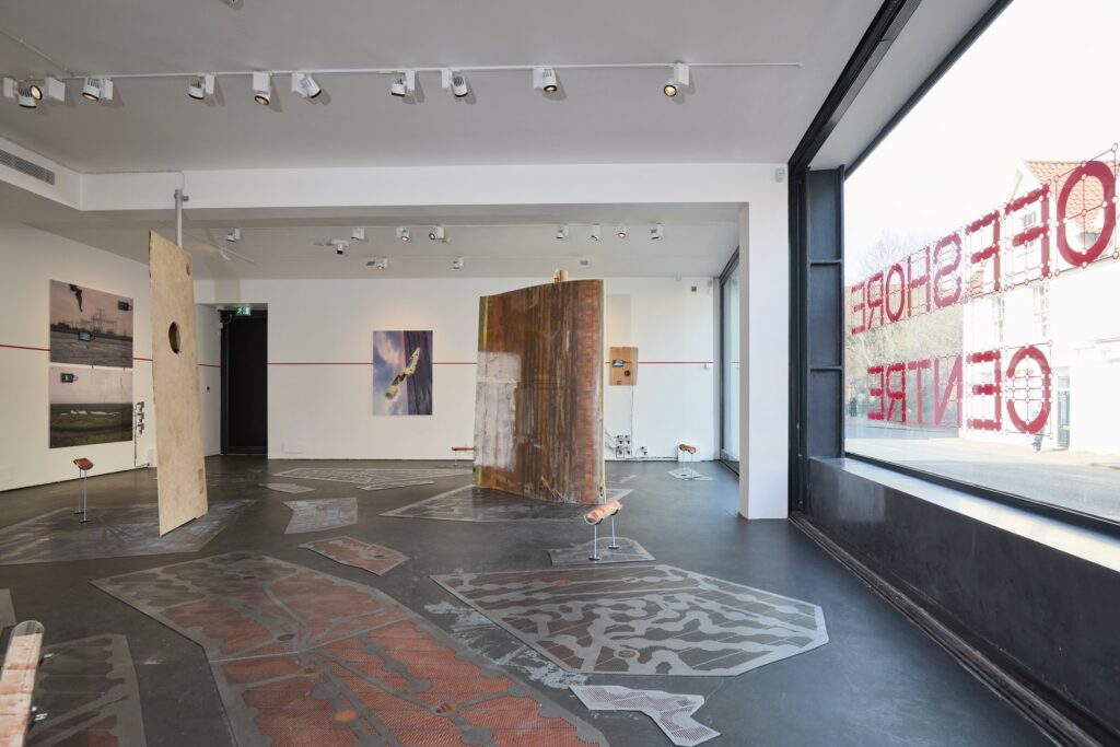A contemporary art gallery with artworks on the walls and large abstract panels suspended in the center. Daylight streams through big windows with red text reading "OFFSHORE CENTER." Patterns decorate the black floor.