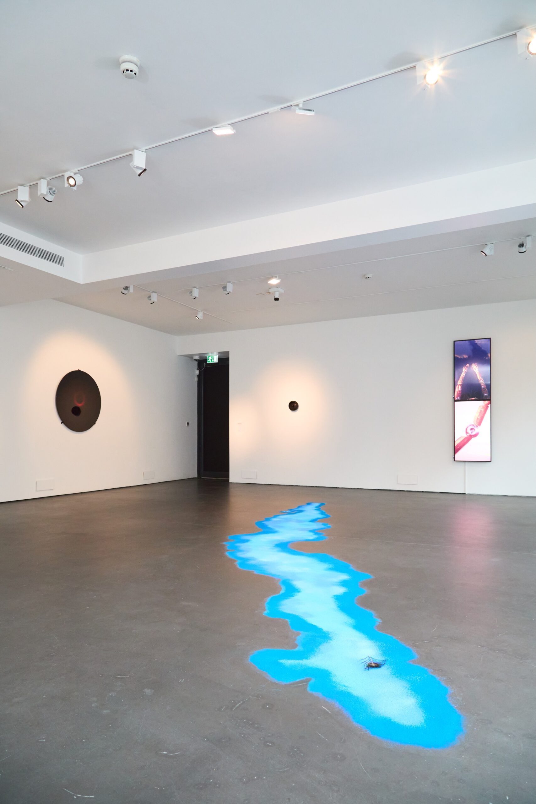 A modern art gallery with white walls features a blue, winding river-like installation on the gray floor. Two circular artworks hang on the walls, and a vertical digital screen displays abstract images. Ceiling lights illuminate the scene.