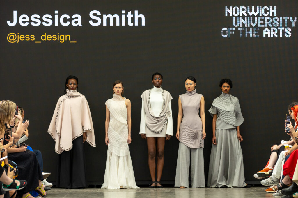 Five models stand on a runway wearing modern, neutral-toned outfits.