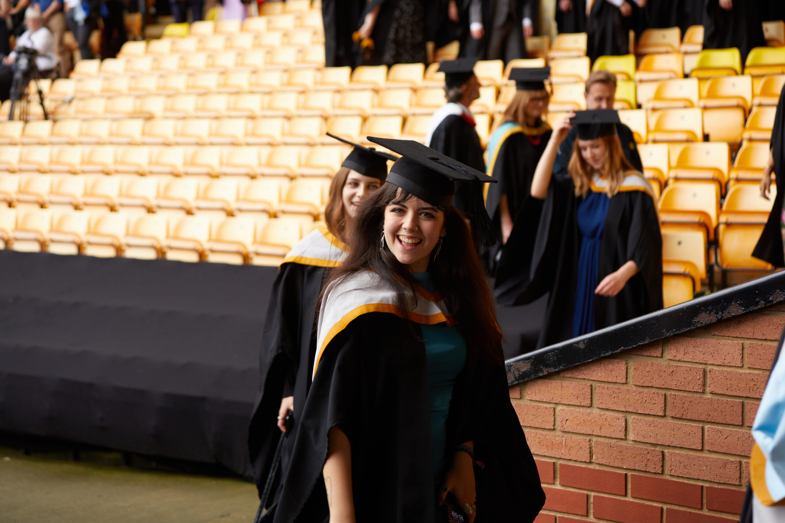 A smiling graduate in cap and gown walks in front of other graduates at a ceremony in a stadium with yellow seats.