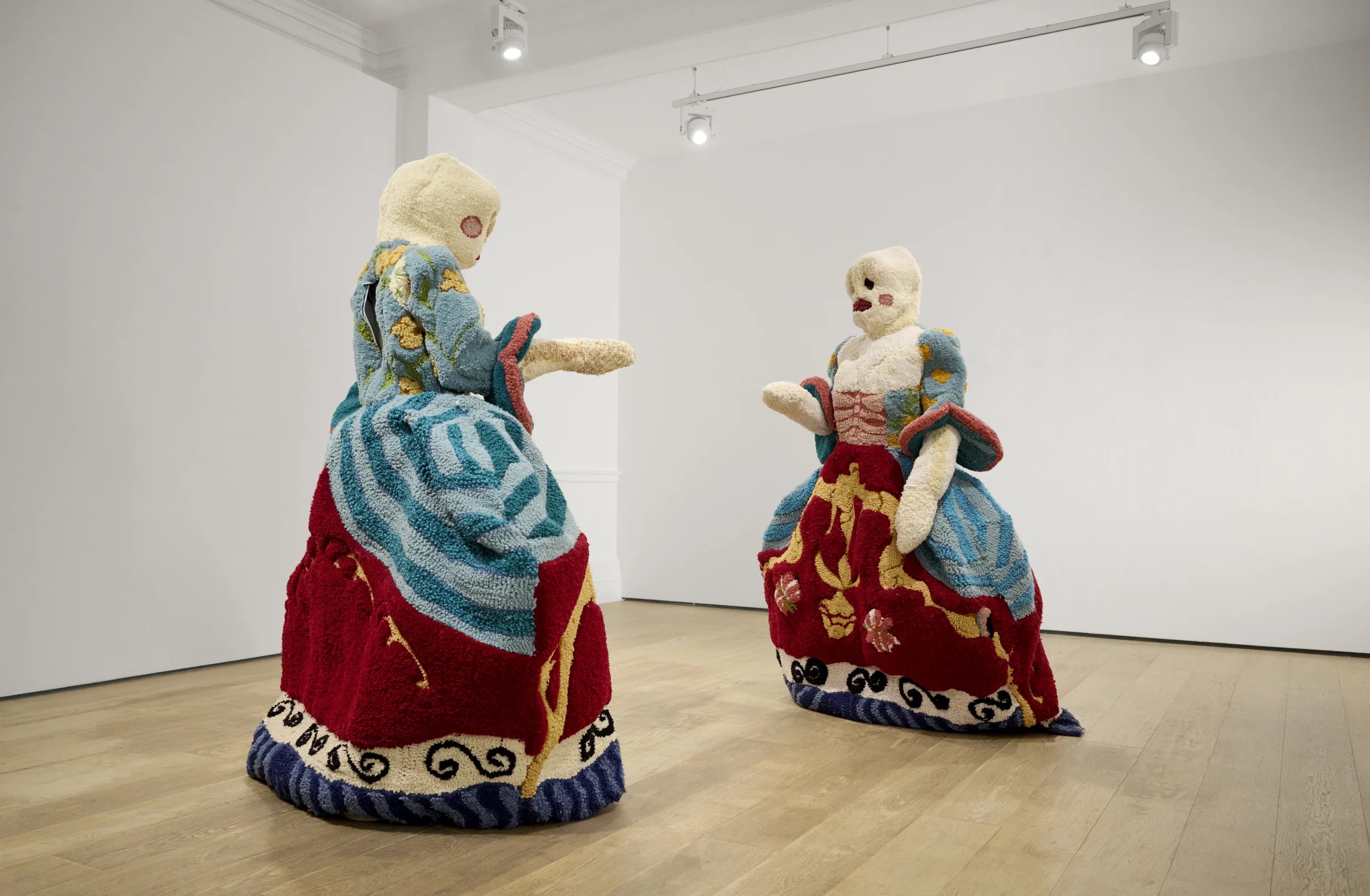 Two large, plush sculptures resembling human figures in colorful, ornate dresses, with simplified faces, stand facing each other in a well-lit art gallery with wooden floors and white walls.