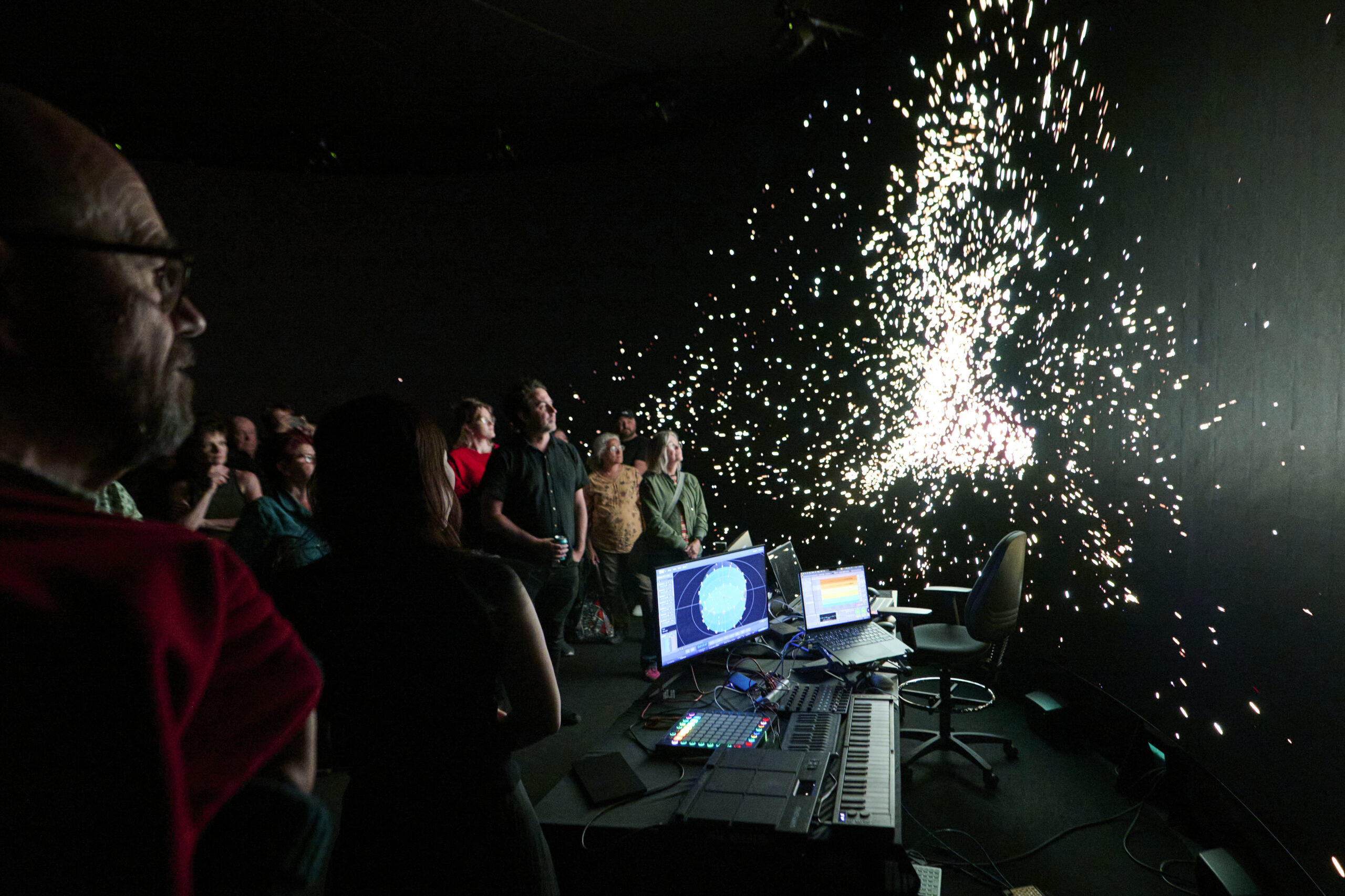 A group of people watch a multimedia performance in a dark room with bright, spark-like visuals on a screen. Electronic music equipment and monitors are set up on a table in the foreground.