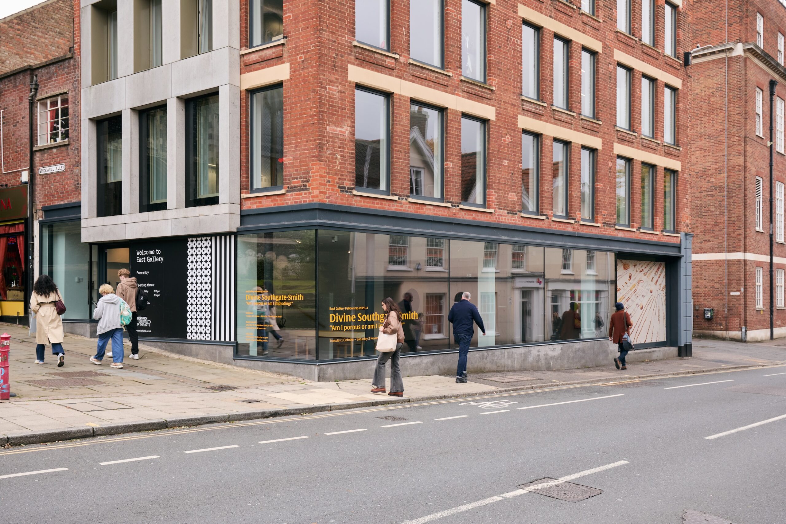 People walk past a modern art gallery with large glass windows on a street corner in a city. The building has a mix of brick and concrete, and exhibition information is visible on the window.
