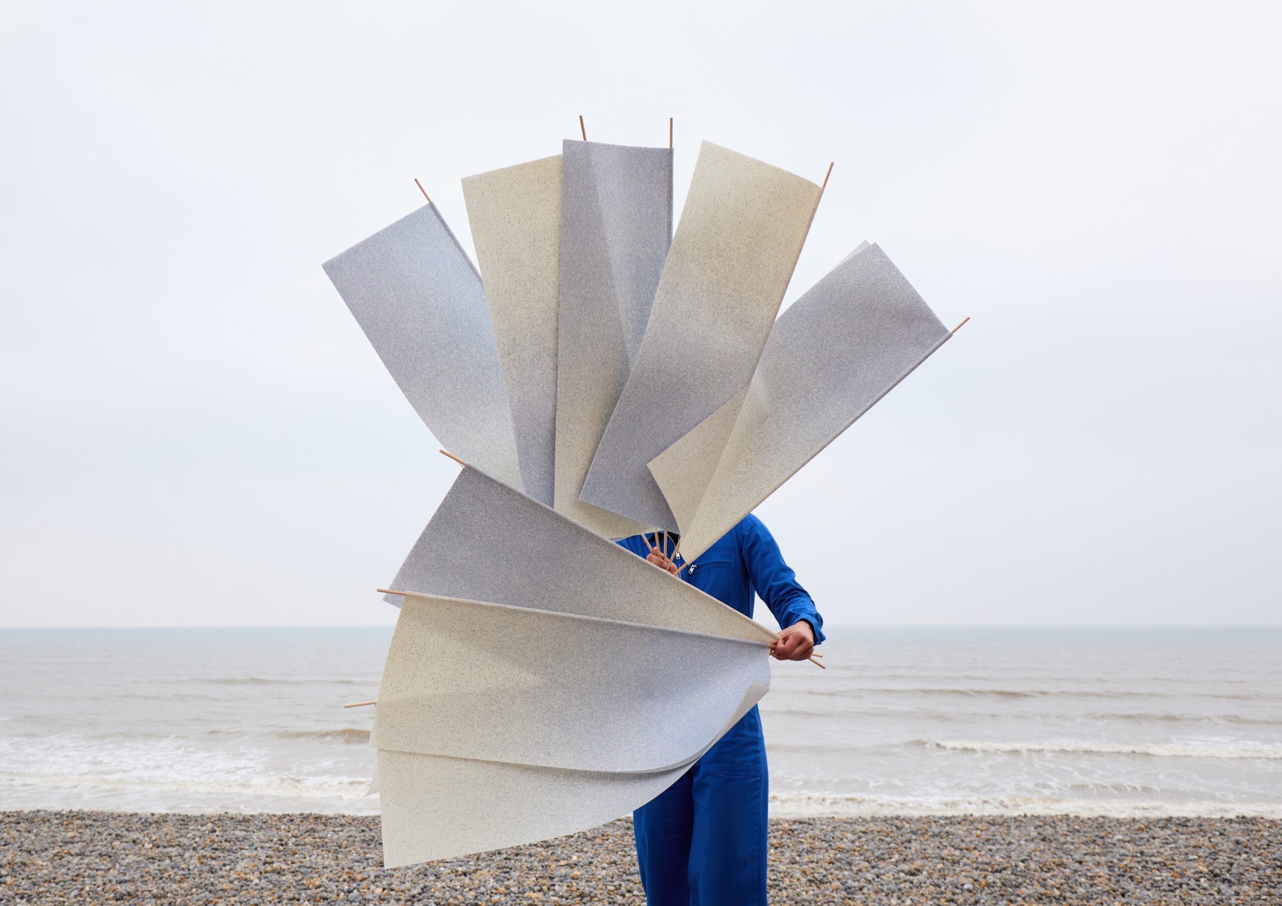 A person in a blue outfit stands on a pebbly beach, holding a large abstract structure made of angular, gray panels that obscure their face and upper body. The sea and cloudy sky are visible in the background.