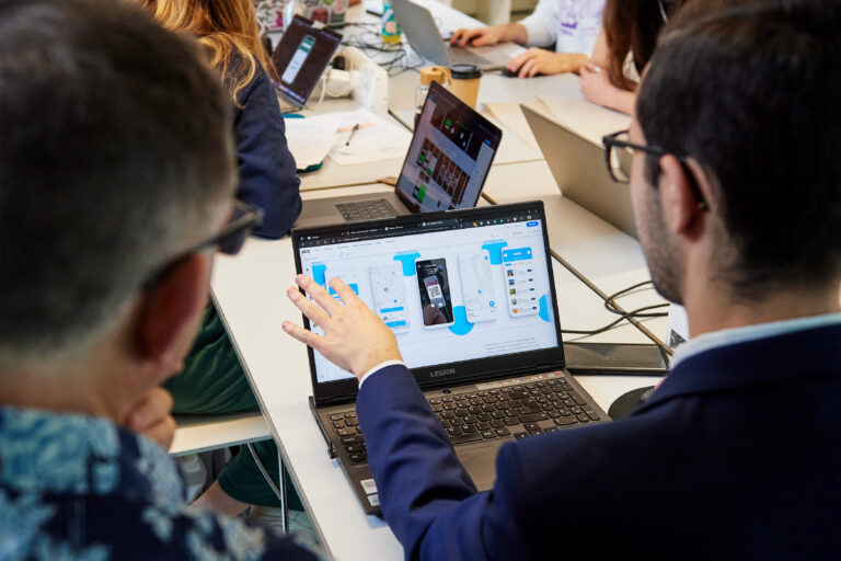 Two people sit at a table with laptops; one points at a laptop screen displaying a UI design for a mobile app while others work in the background. The scene suggests a collaborative work or design meeting.