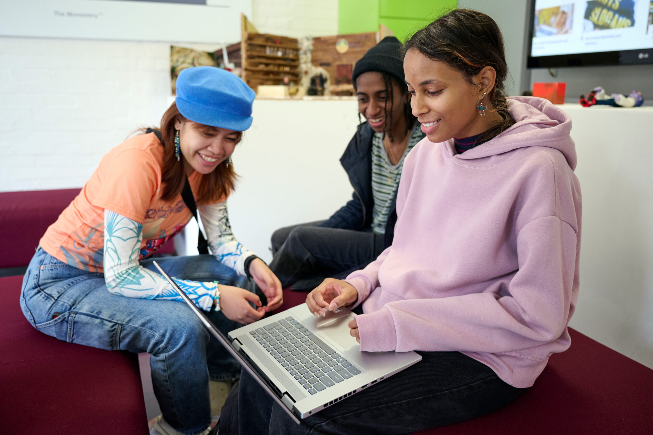 Three young people sit together on a couch, smiling and looking at a laptop. One wears a blue beret, another a black hat, and the third a lavender hoodie. The setting appears to be a casual, modern indoor space.