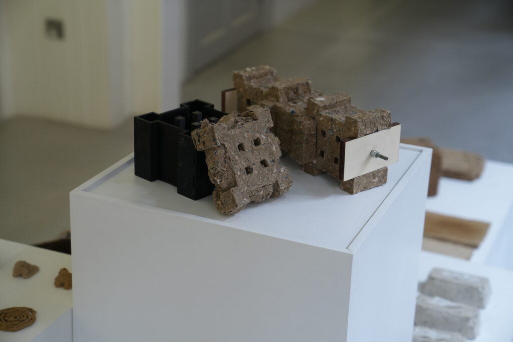 A display of textured, square blocks, some with holes, rests on a white pedestal. One block is held together by a metal plate and bolt, and black rectangular moulds are also visible behind the blocks.