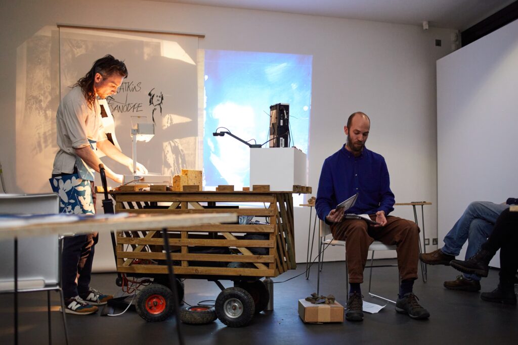 A person stands at a table with papers and equipment, illuminated by a projector. Another person sits nearby reading, with a wooden cart and boxes between them in a modern, minimal room.