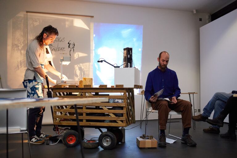 A person stands at a table with papers and equipment, illuminated by a projector. Another person sits nearby reading, with a wooden cart and boxes between them in a modern, minimal room.