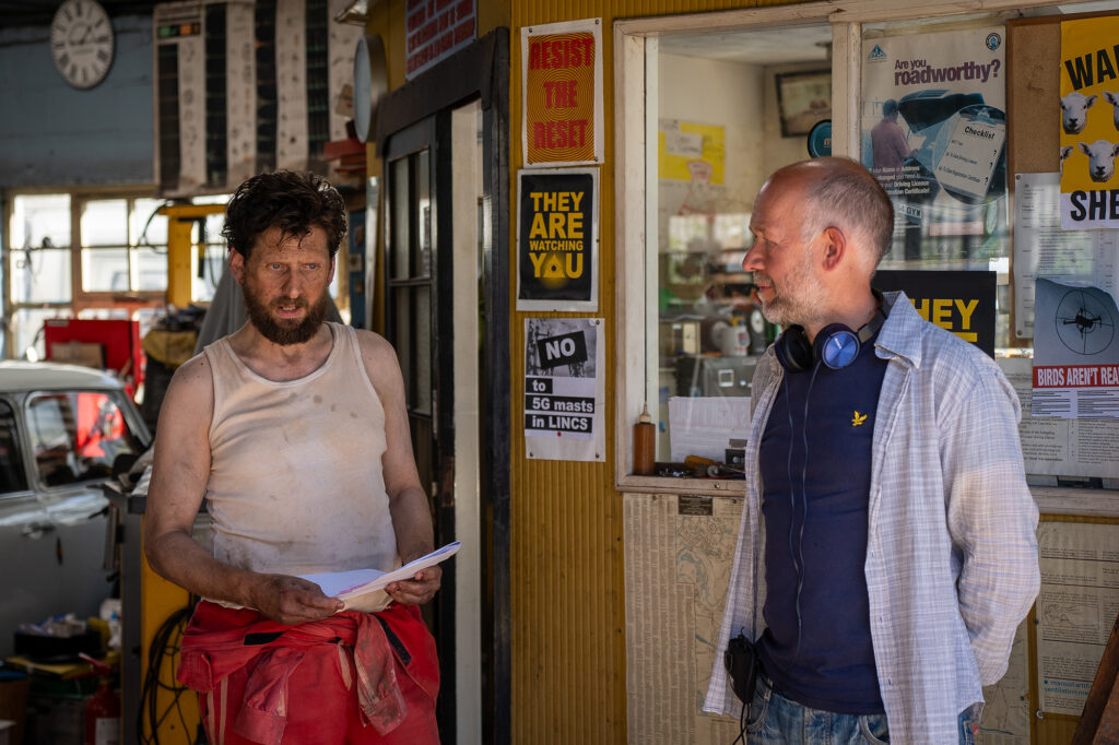 Two men stand in a messy auto repair shop. One, in a dirty tank top, holds papers and looks disheveled. The other, in a checked shirt with headphones around his neck, listens. Signs and papers cover the office wall behind them.
