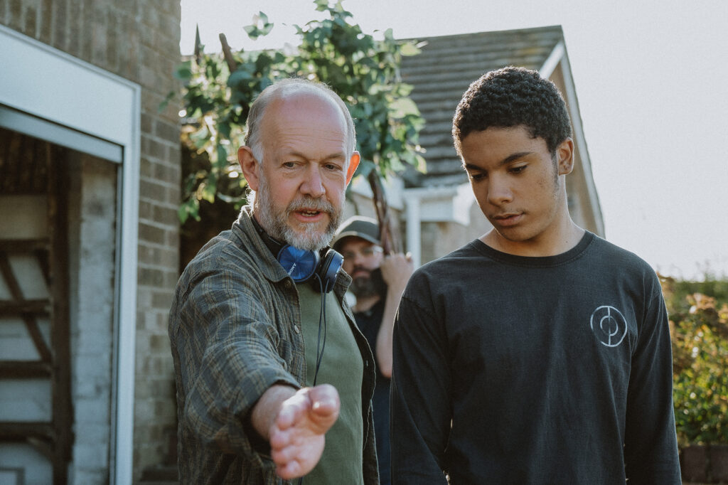 A man with headphones around his neck gestures with his hand while speaking to a young man in a black shirt outdoors, with a third person and greenery in the background.