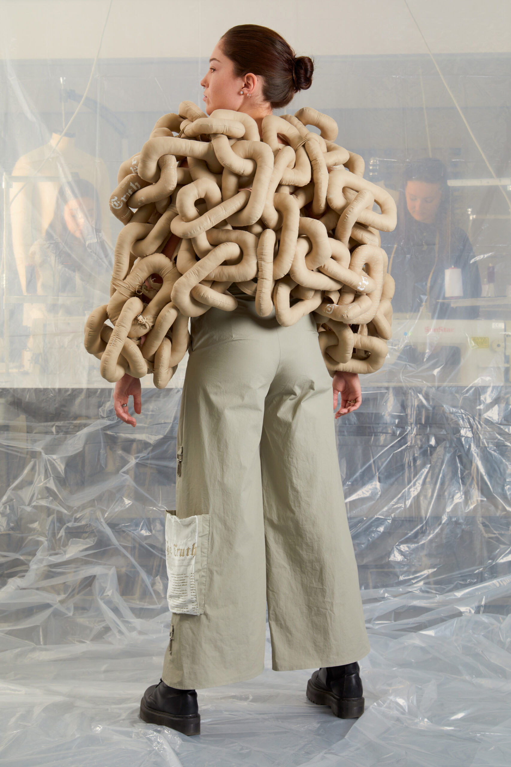 A woman stands with her back to the camera in an industrial clearing, wearing a unique jacket made of large, padded beige tubes over light gray wide-legged pants and black shoes. Plastic sheeting and people are visible in the background.