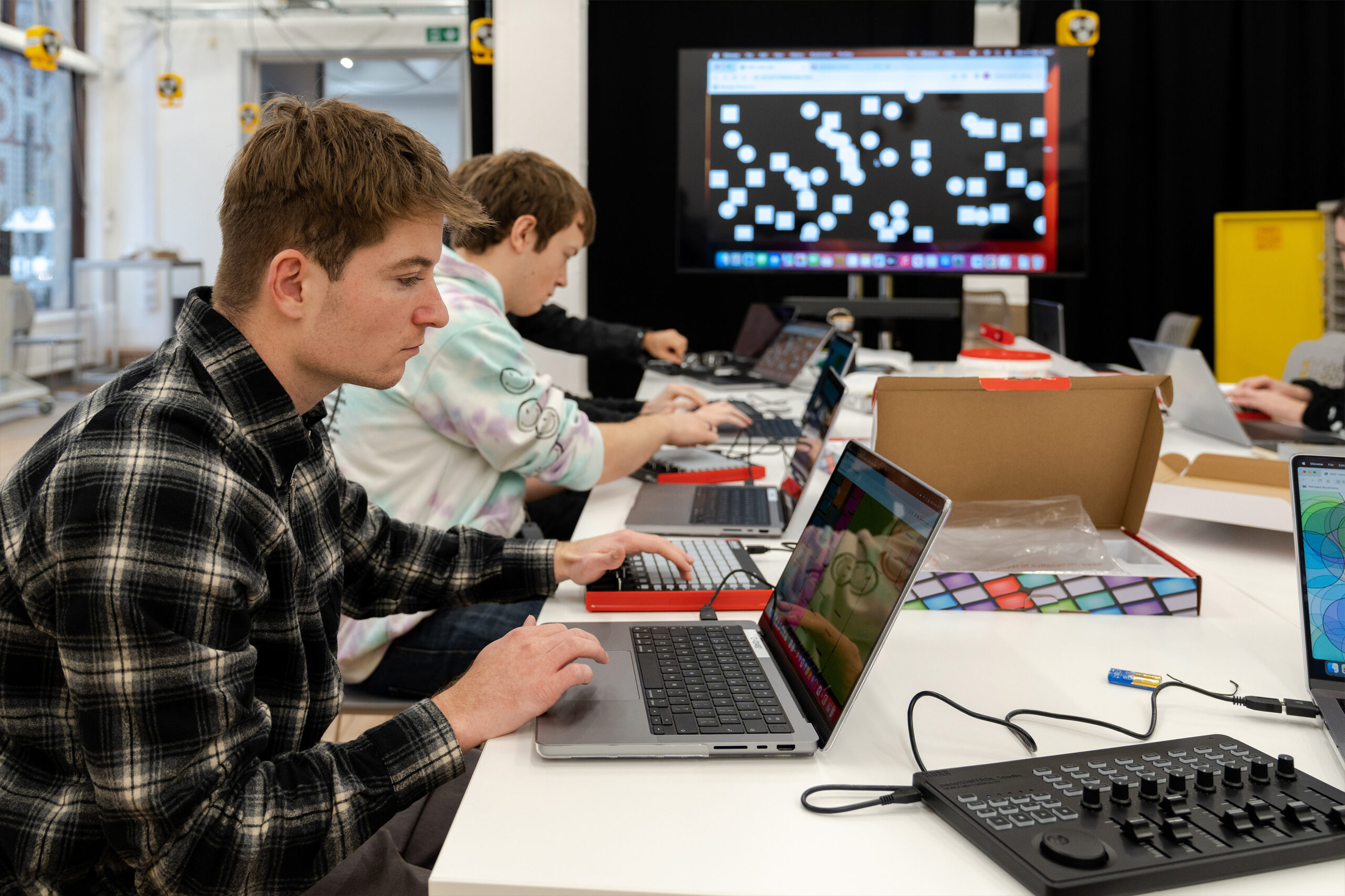 Two people work on laptops at a table with electronic equipment and colorful circuit boards. A large screen in the background displays a pattern of white squares on a black background.