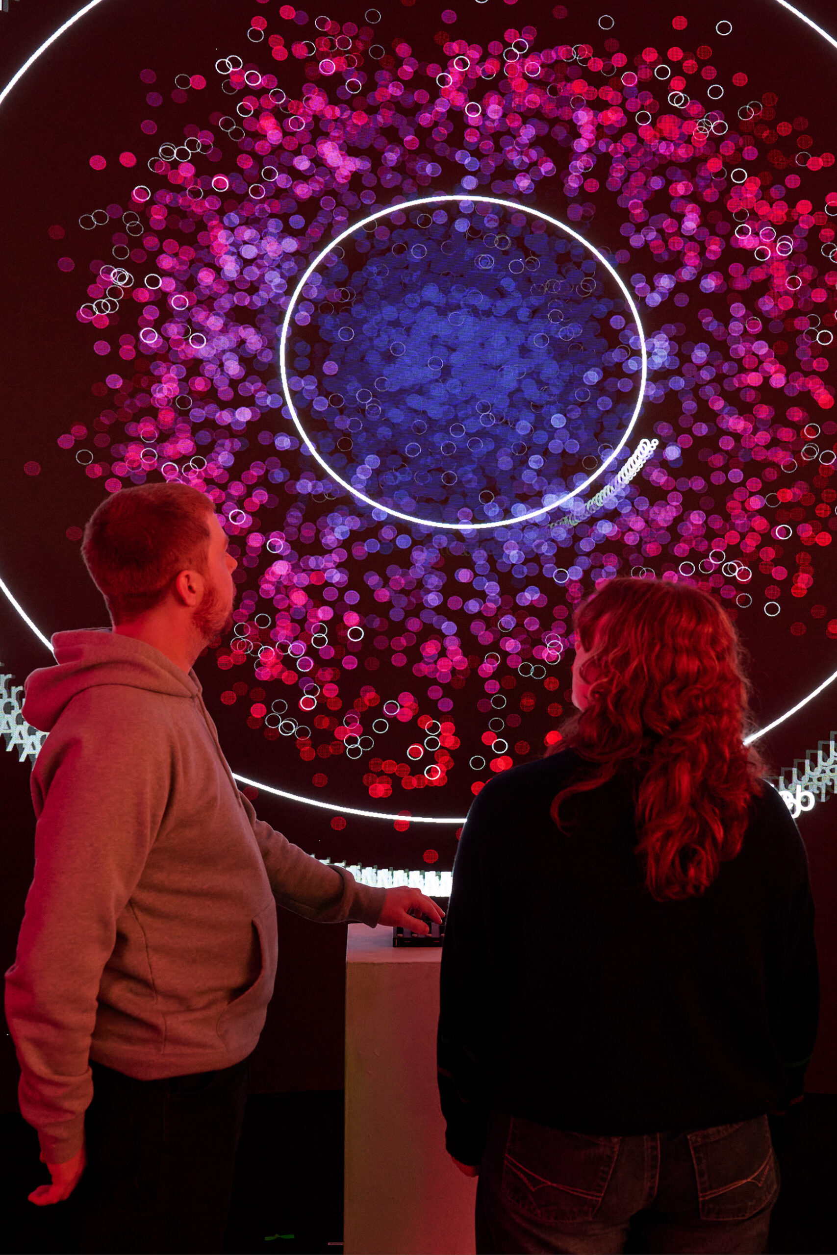 Two people stand facing an interactive digital art display with swirling blue and pink lights and circular patterns on a dark clearing. The vibrant design creates a dynamic, futuristic atmosphere.