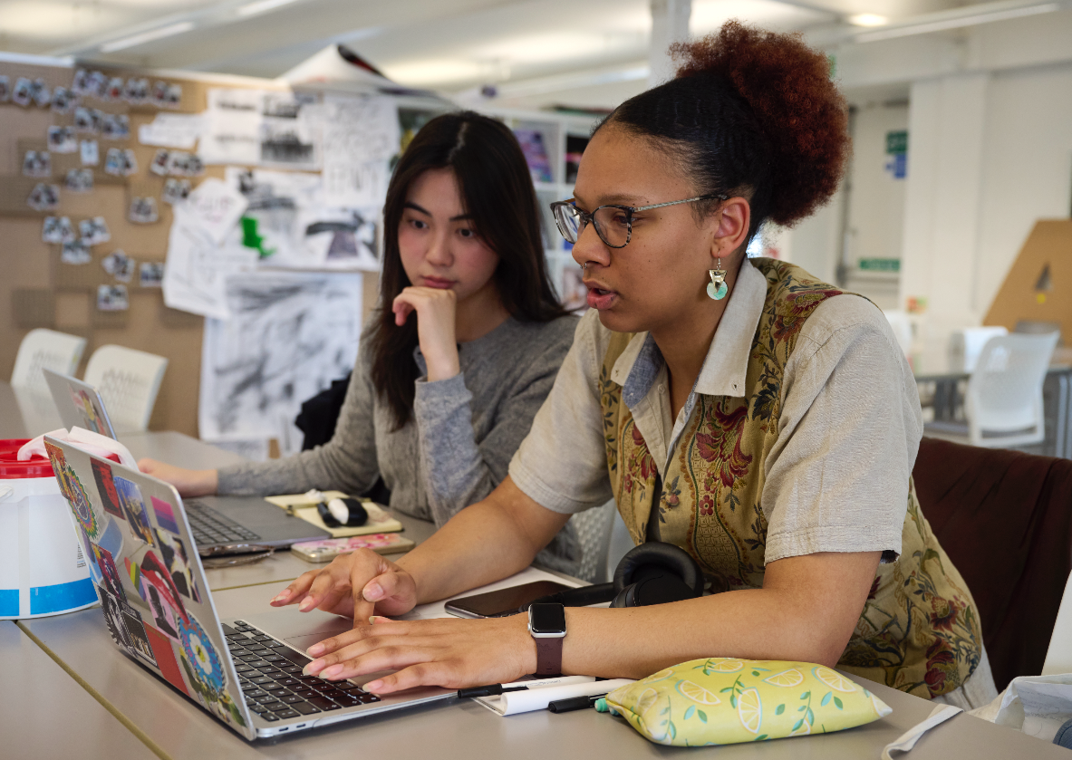 Two focused students sit at a table working on laptops. One types while the other observes. The background features a bulletin board with photos and papers, and the workspace is bright and organized.