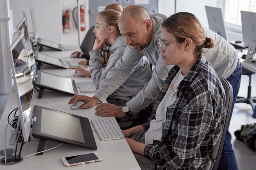 Three people work at computers in a bright classroom. A man leans over to assist a student, while another student sits nearby focused on her screen. A smartphone lies on the desk in the foreground.