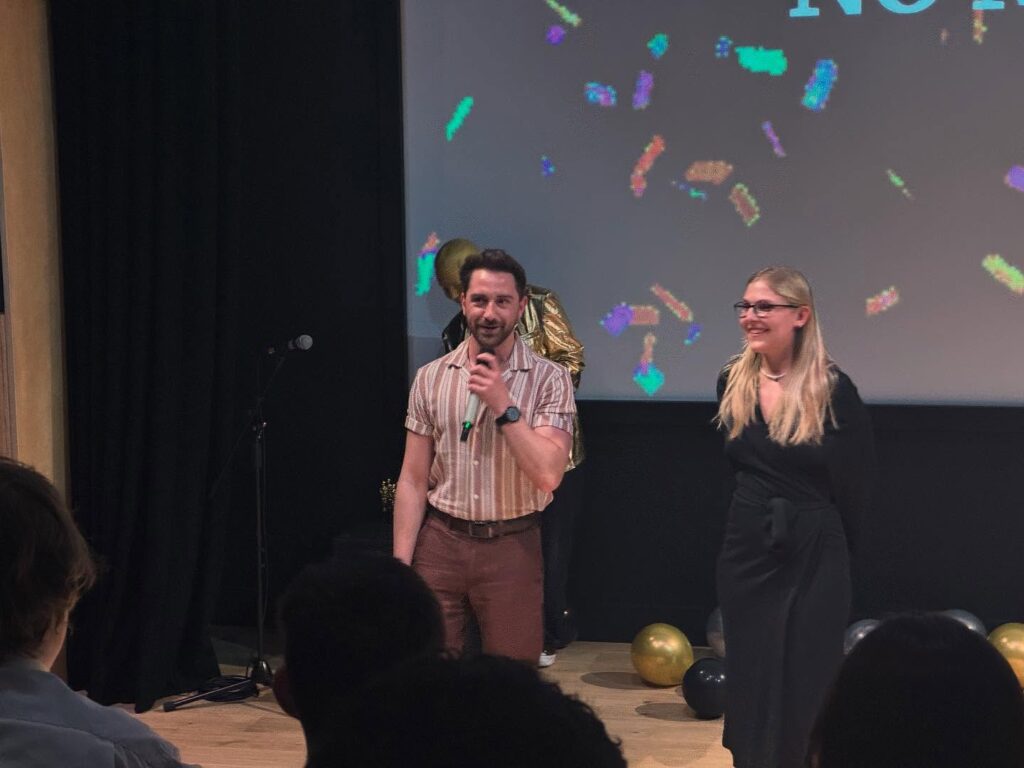 A man holding a microphone and a woman stand on stage, smiling at an audience. Colorful confetti graphics are projected behind them, along with gold and black balloons on the floor.