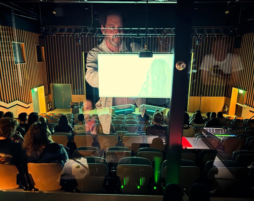 A small theater with rows of seats facing a screen showing a person; a man’s reflection appears on the glass, overlapping the scene. Several audience members are seated, watching attentively.