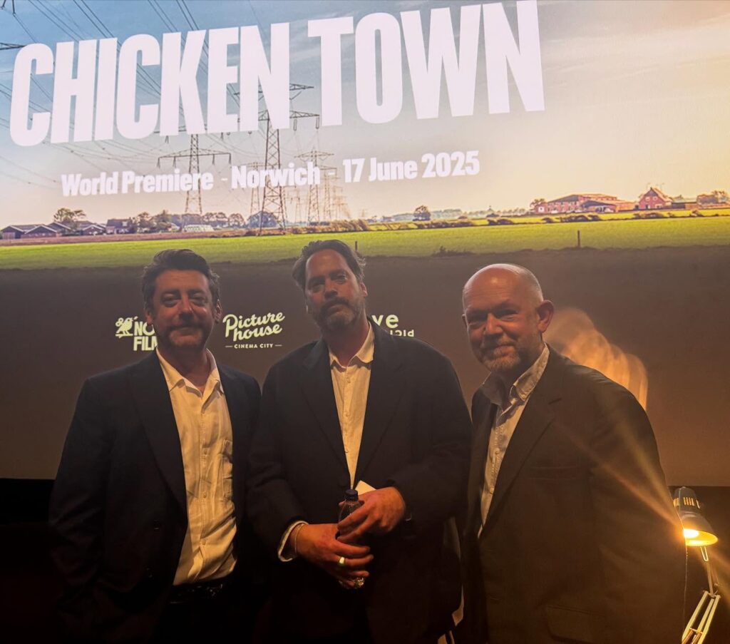 Three men in suits stand in front of a large screen displaying “CHICKEN TOWN,” with details about a world premiere in Norwich on 17 June 2025. The background image shows a rural scene with power lines and houses.