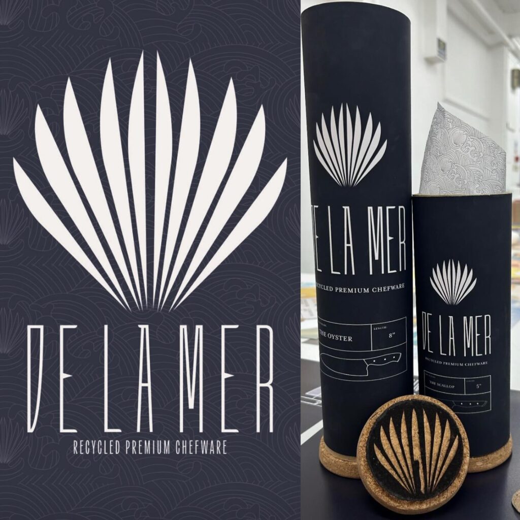 A display of De La Mer recycled premium chefware packaging, featuring a cylindrical black container with a white stylized leaf logo, decorative paper, and two round cork items.