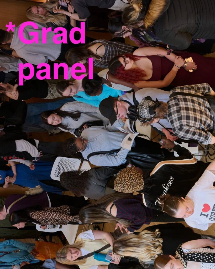 A crowded room of students, some seated and some standing, gathered around tables for a grad panel event. Large pink text at the top left reads “*Grad panel.”.