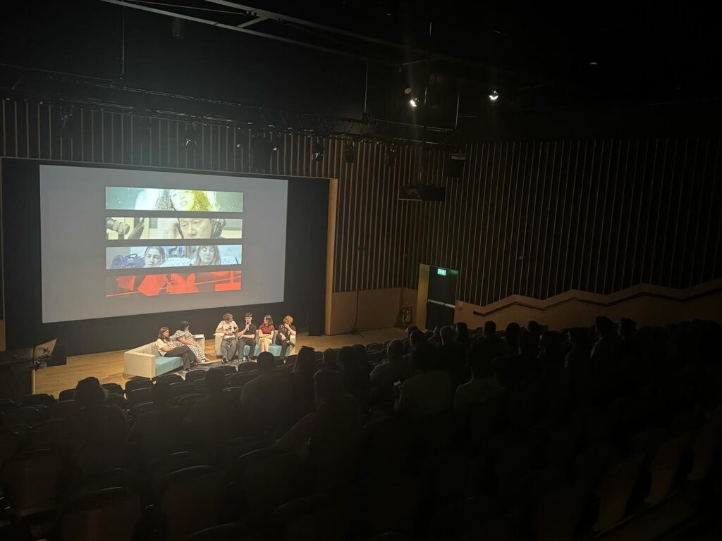 A panel of six people sits on a stage in front of a large screen displaying images, addressing an audience in a dimly lit auditorium.
