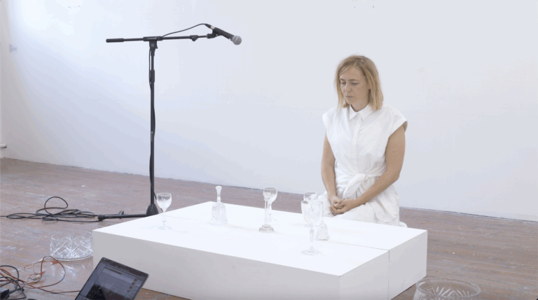 A woman in a white dress kneels beside a white platform with several glass goblets on it. A microphone on a stand is positioned above the glasses. The scene is set in a minimal, white-walled room with wooden floors.