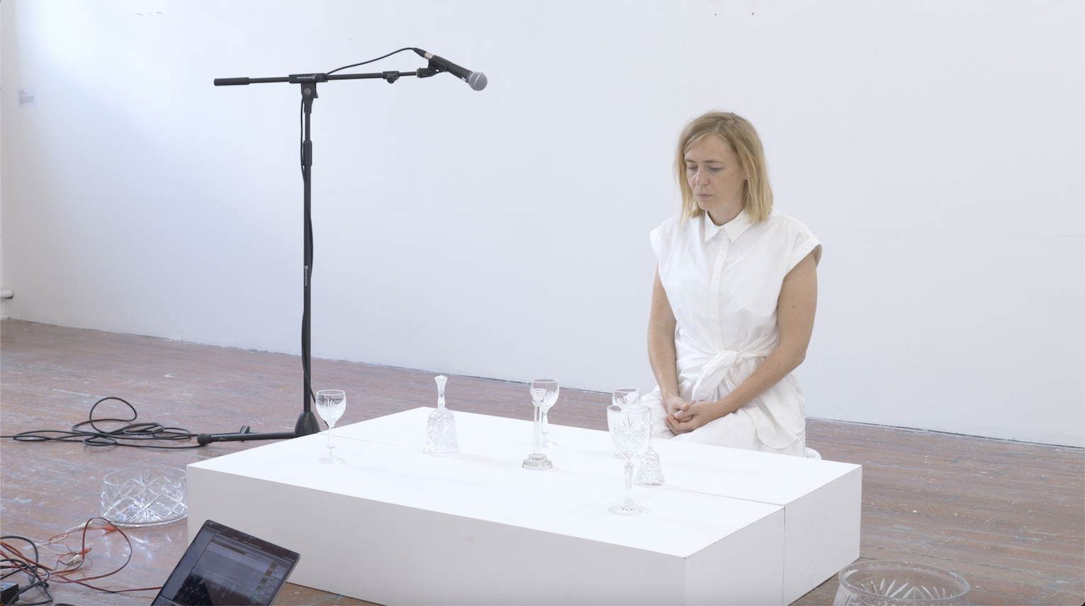 A woman in a white dress kneels beside a white platform with several glass goblets on it. A microphone on a stand is positioned above the glasses. The scene is set in a minimal, white-walled room with wooden floors.