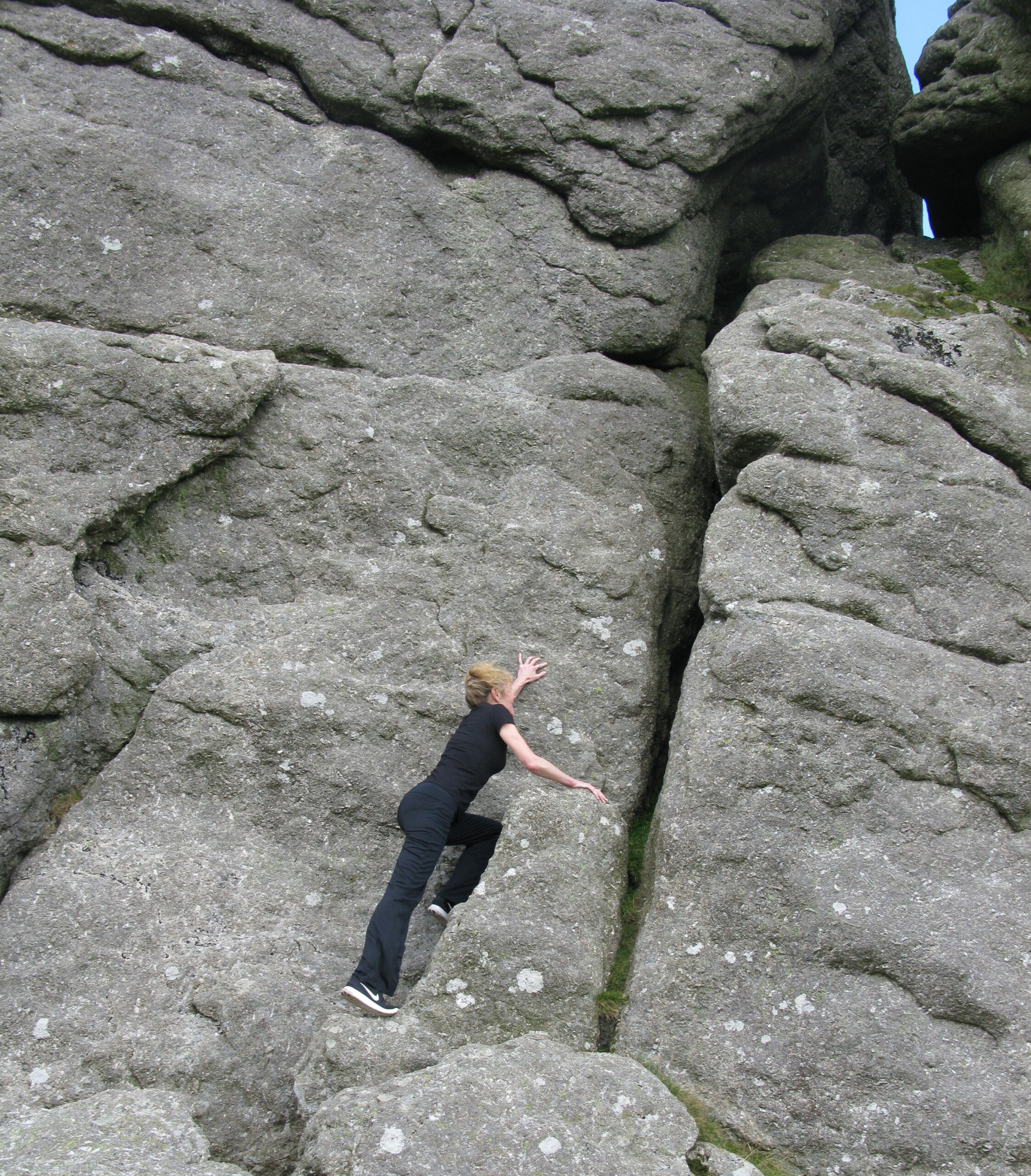 A person dressed in black climbs a steep, rugged rock face, reaching upward with one hand while bracing themselves with their feet and other hand in a narrow crevice.