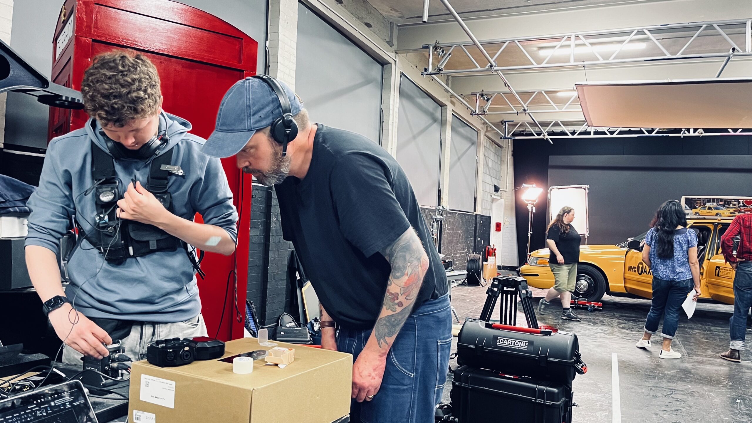 Two crew members wearing headsets work with equipment on set. Behind them, a film scene with a yellow taxi, lights, and people in motion unfolds in a spacious industrial studio.