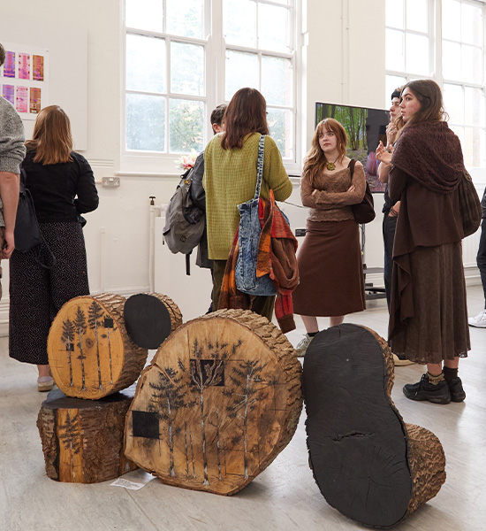 People gather in a bright gallery space with large windows, talking and observing art. Tree stump sculptures with nature designs are displayed in the foreground at the Norfolk Art and Design Competition Exhibition. Some people view art on the walls.