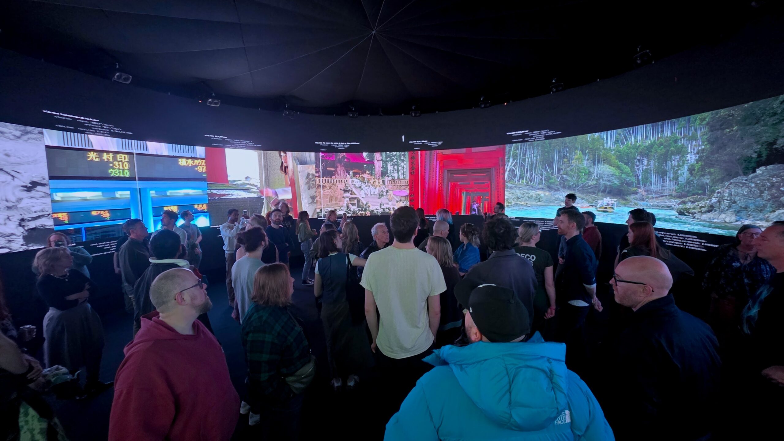 A group of people stand inside a dark circular room surrounded by large, colorful, illuminated screens displaying scenic images and text, creating an immersive exhibition environment enhanced by creative technologies.