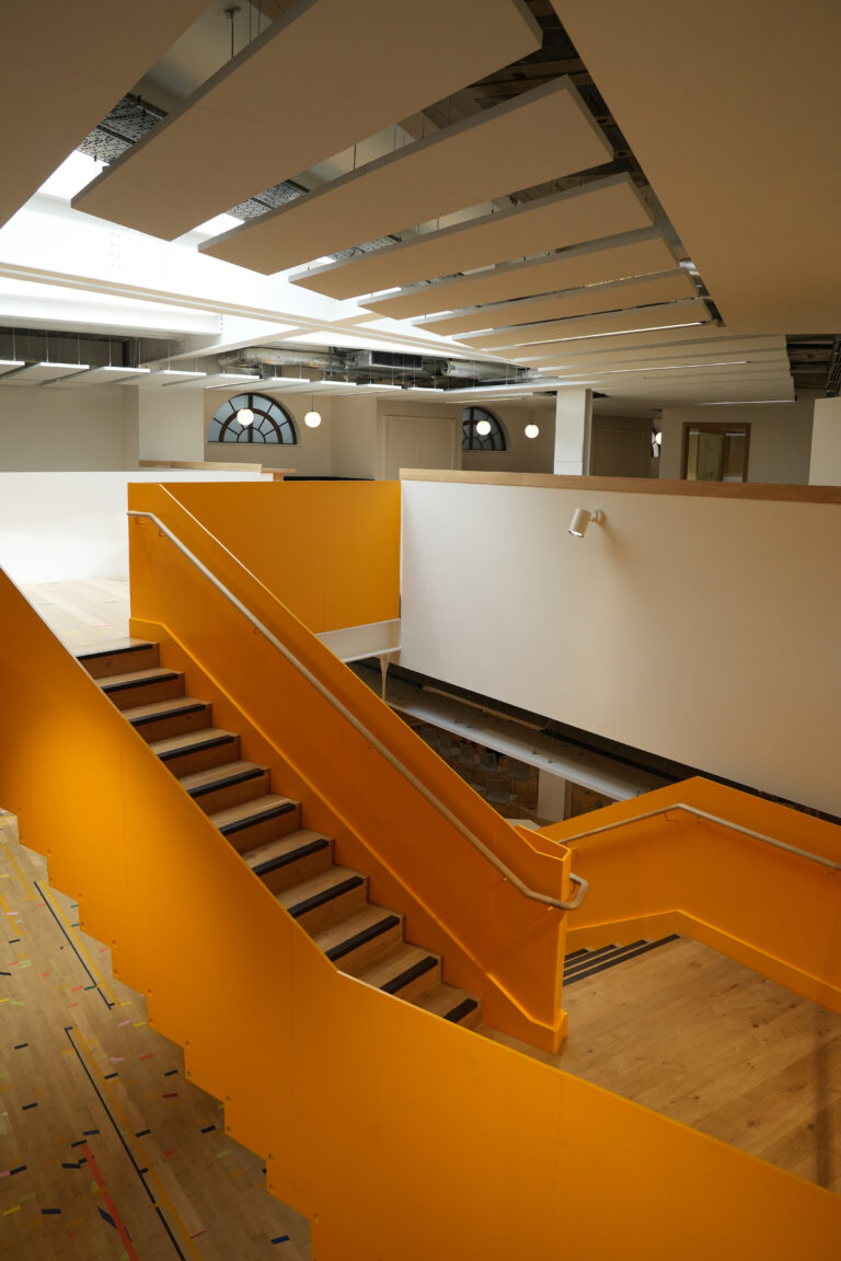 A modern interior space with a bright yellow staircase, wooden floors, white walls, and large windows with rounded tops.