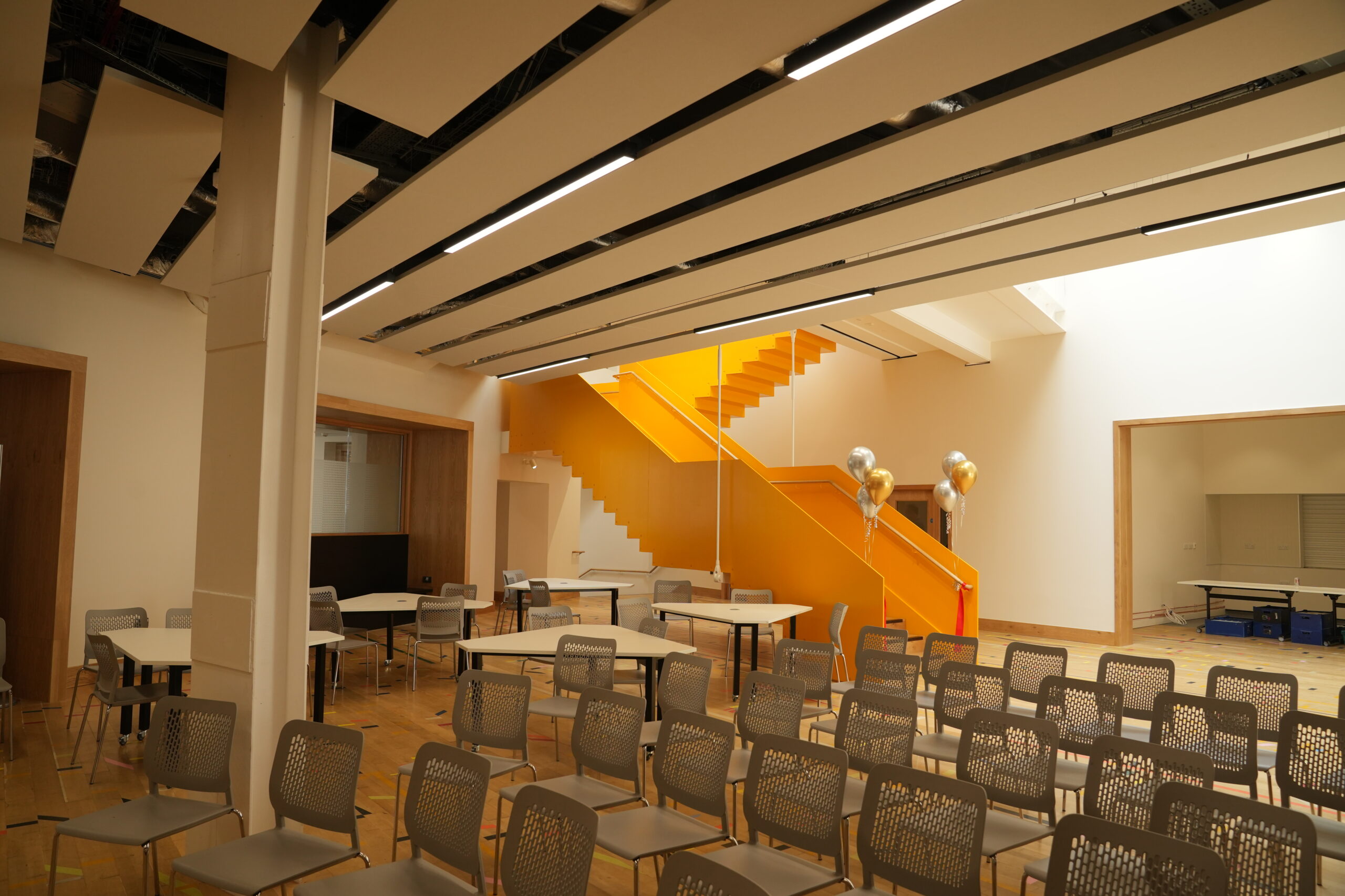 A modern room with rows of chairs and tables and a bright orange staircase. Balloons are tied near the stairs, and the ceiling shows exposed panels.