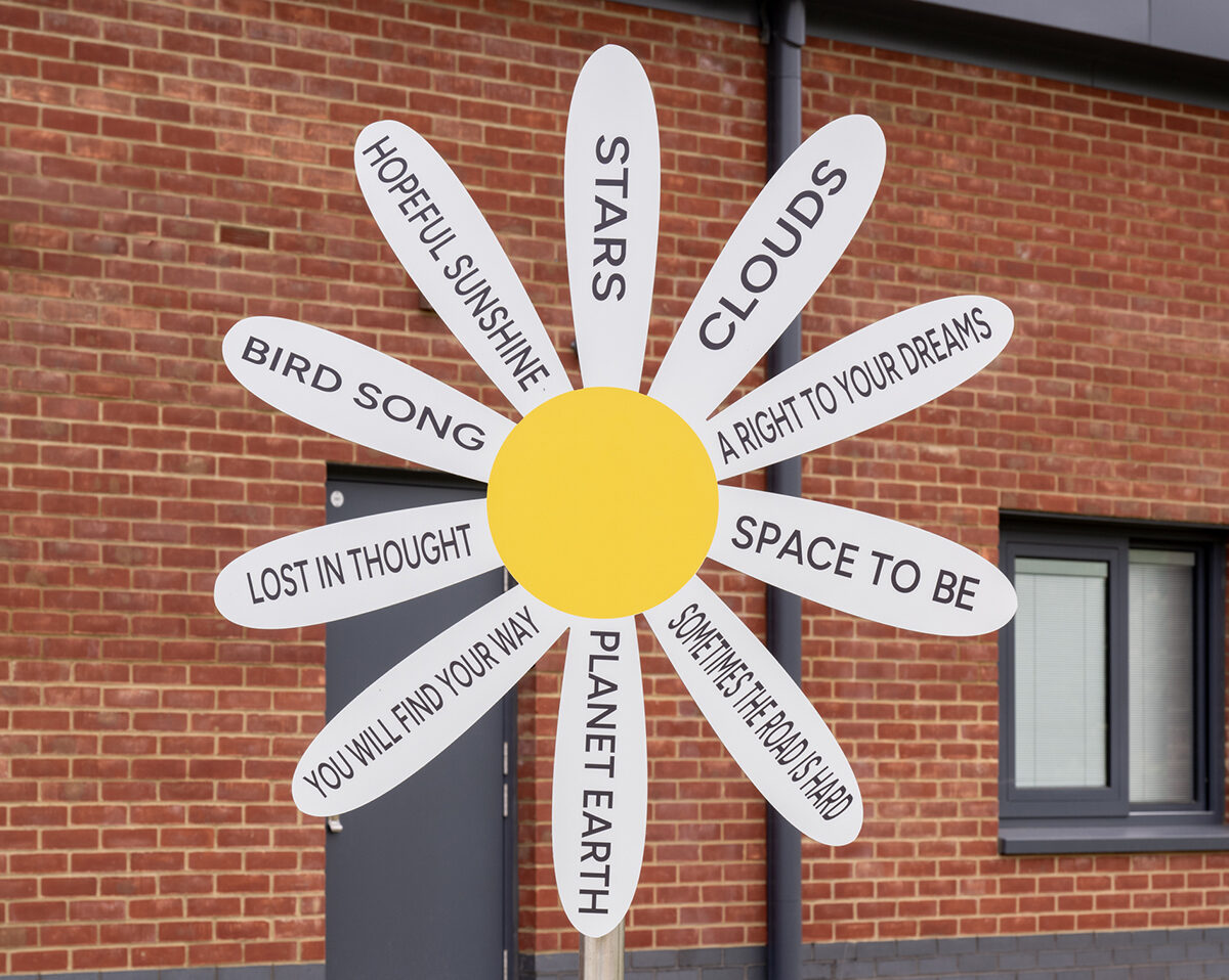 A daisy-shaped sign with white petals, each labeled with phrases like “STARS,” “CLOUDS,” “SPACE TO BE,” and “YOU WILL FIND YOUR WAY,” stands in front of a red brick building, celebrating creative arts and health.