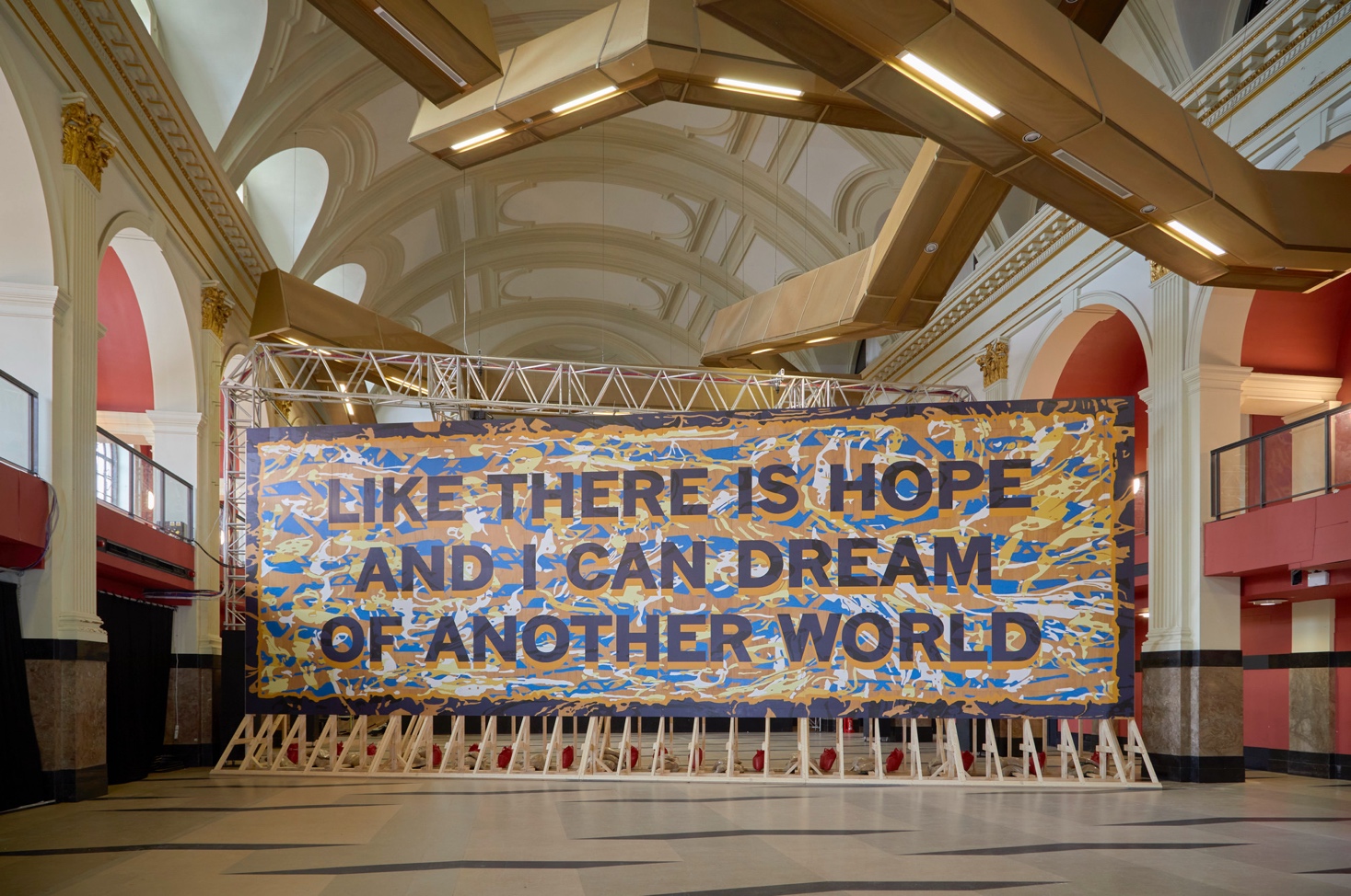 A large, colorful banner in a grand hall reads, "LIKE THERE IS HOPE AND I CAN DREAM OF ANOTHER WORLD." Inspired by vibrant murals, the banner is decorated with abstract patterns and supported by a metal frame.