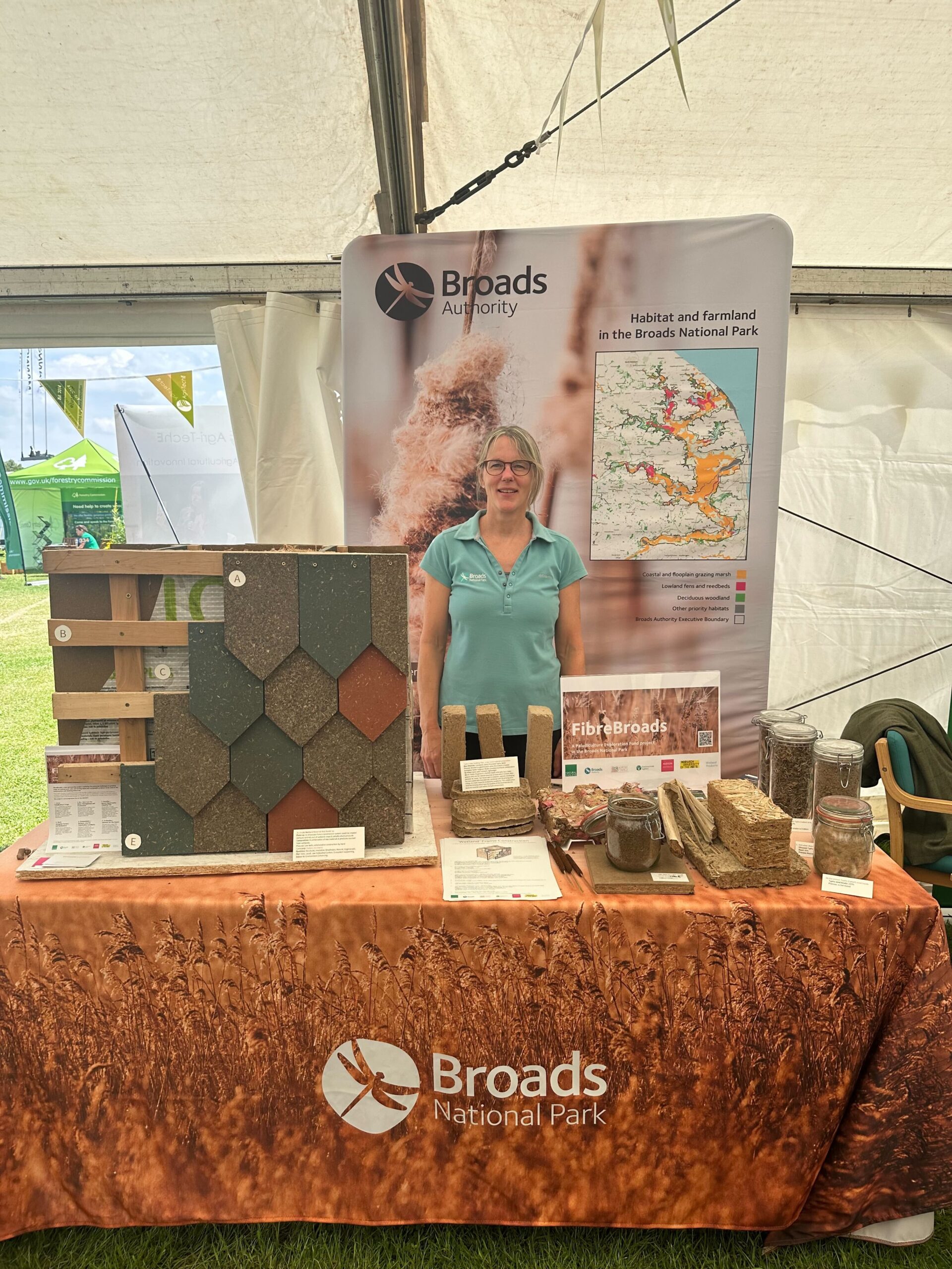 A woman stands at a Broads National Park display booth featuring paludiculture building materials, jars of samples, informational pamphlets, and a map poster inside a tent at an outdoor event.