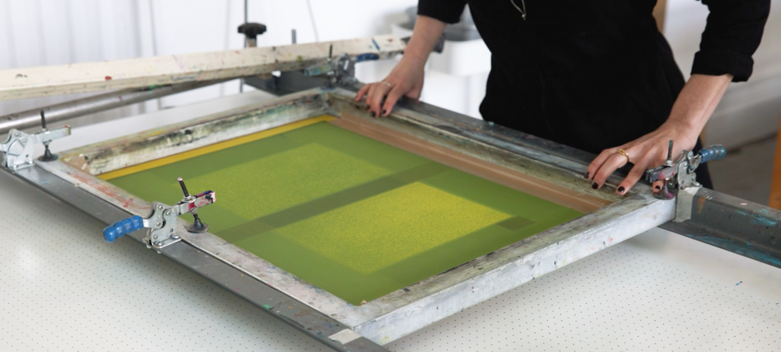 A person operates a screen printing press in a print workshop, holding the frame steady as yellow-green ink spreads over the screen. The setup rests on a white, perforated table in a well-lit workspace.