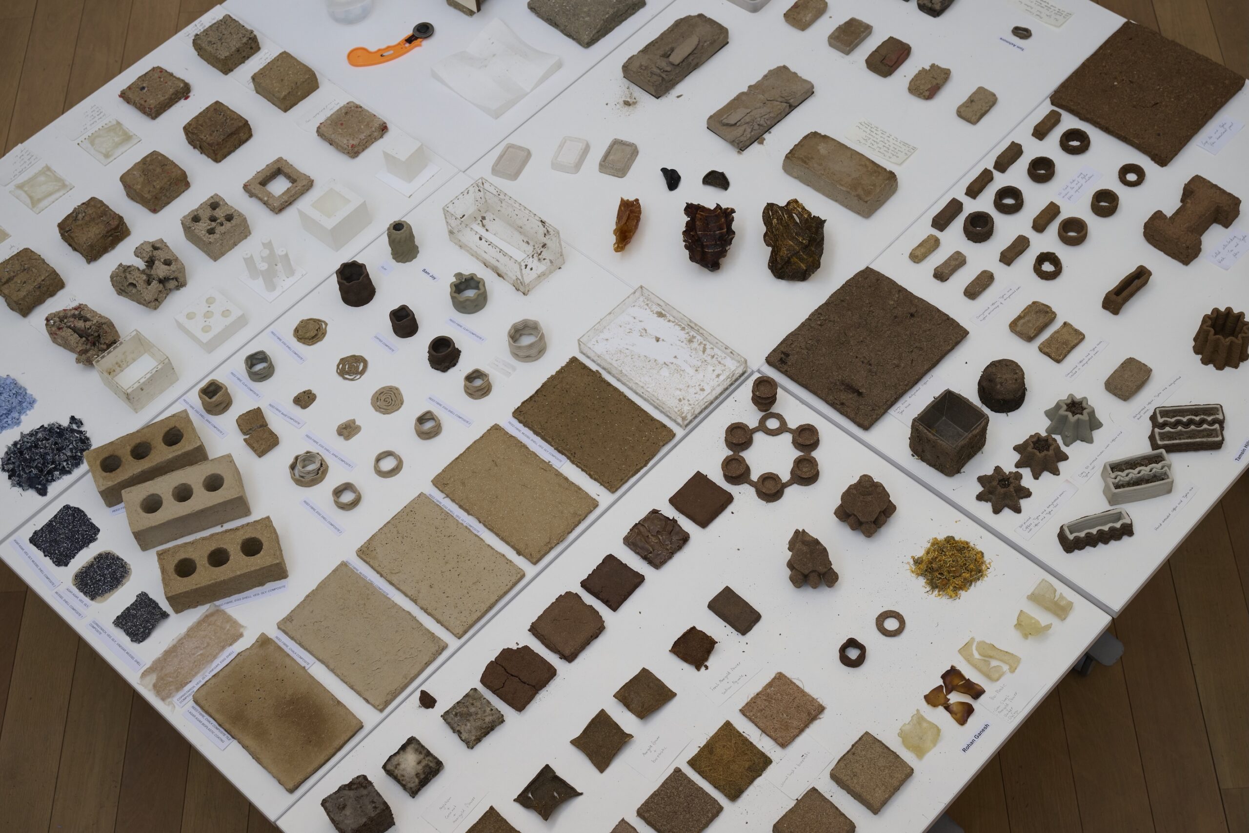 A display of various natural and man-made material samples, including paludiculture-derived products, as well as different shapes and colors of tiles, bricks, powders, blocks, and geometric forms, arranged neatly on white tables.