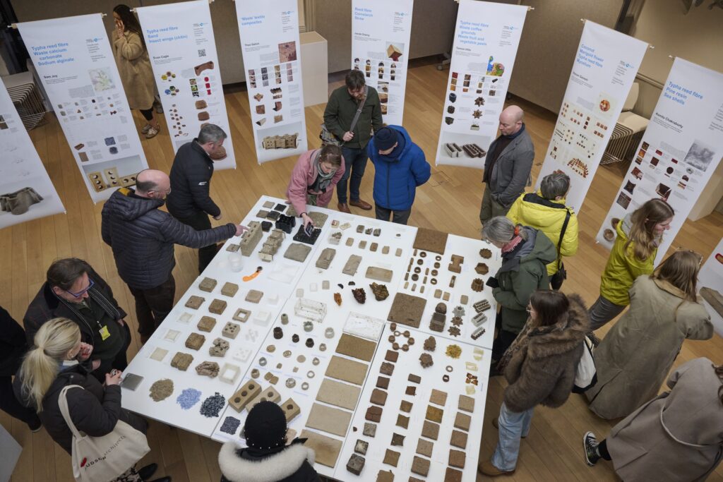 People stand around and examine tables displaying various small natural material samples related to paludiculture in an exhibition space; informational posters with images and text are displayed in the background.