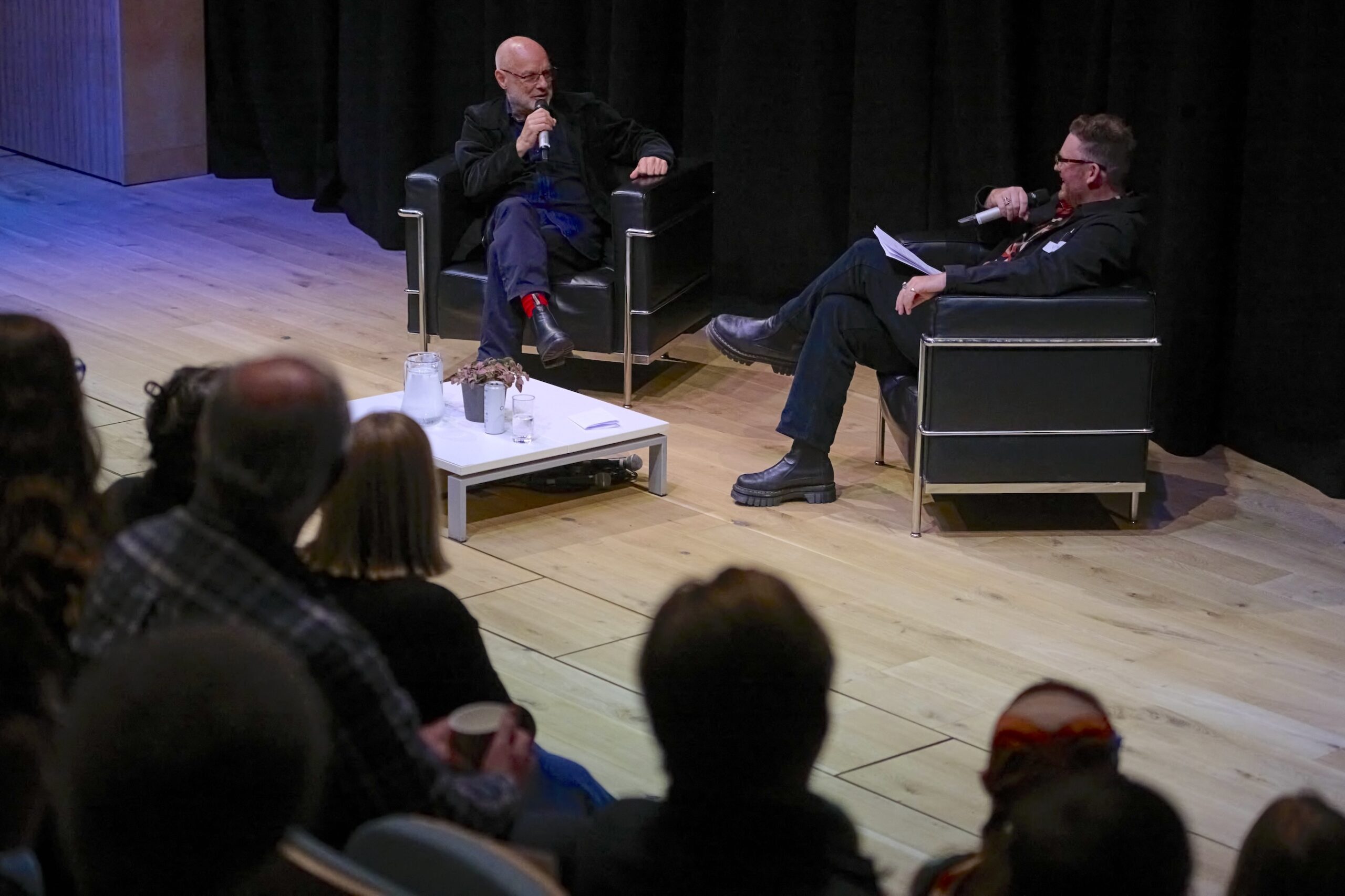 Two people sit in armchairs on a stage, having a talk with microphones as the audience watches. A small table with drinks is between them, and the floor is light wood.