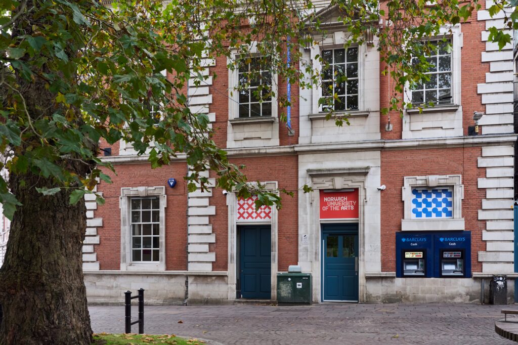A red-brick building with white stone trim, labeled "Norwich University of the Arts." A large leafy tree is in the foreground.