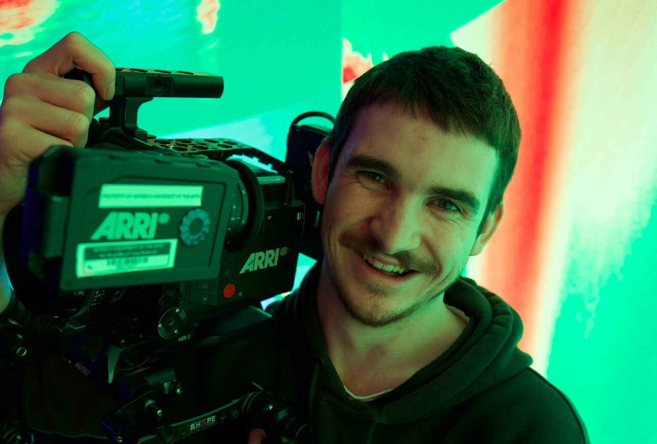 A smiling man holds a large ARRI video camera on his shoulder, standing in front of a brightly lit, colorful green and red background. He wears a dark hoodie and has a tattoo on his forearm.