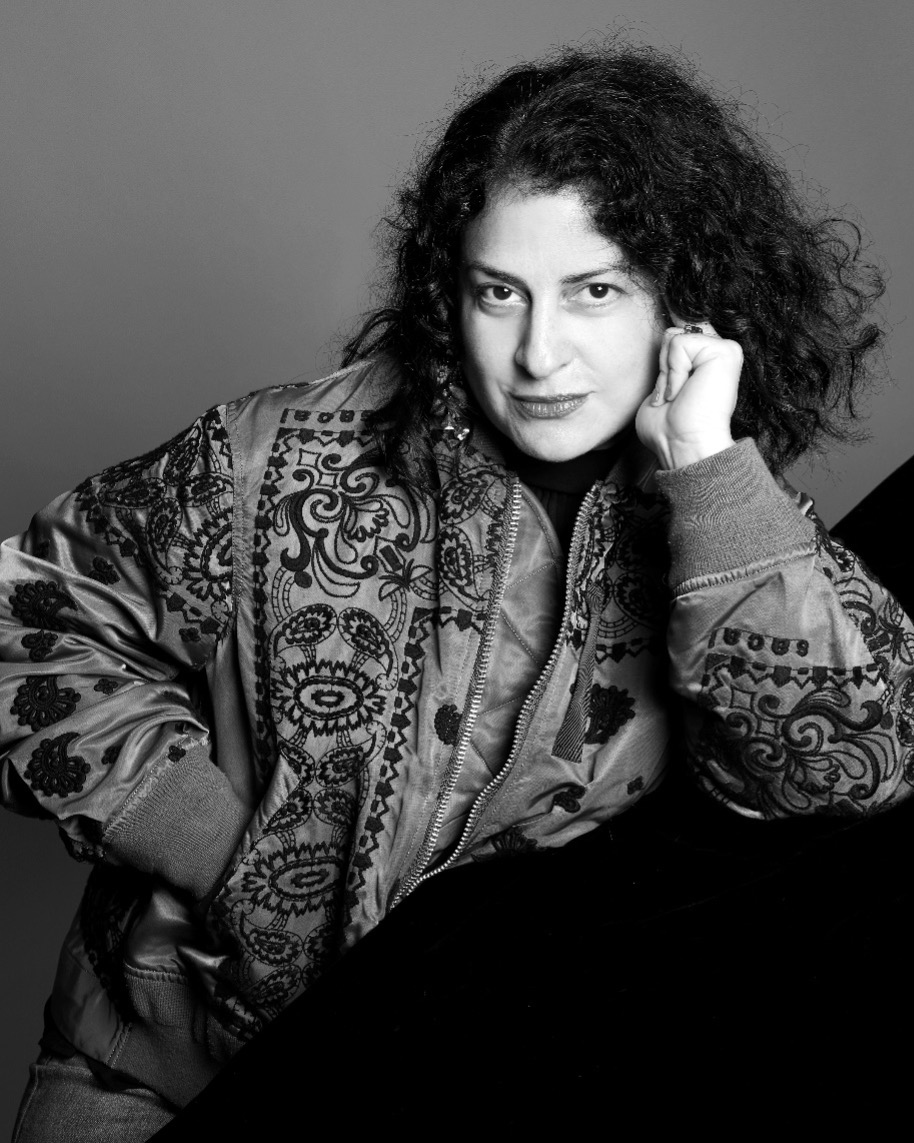 A woman with curly hair wears a patterned jacket and leans against a surface, resting her head on her hand, looking confidently at the camera; the photo is black and white.
