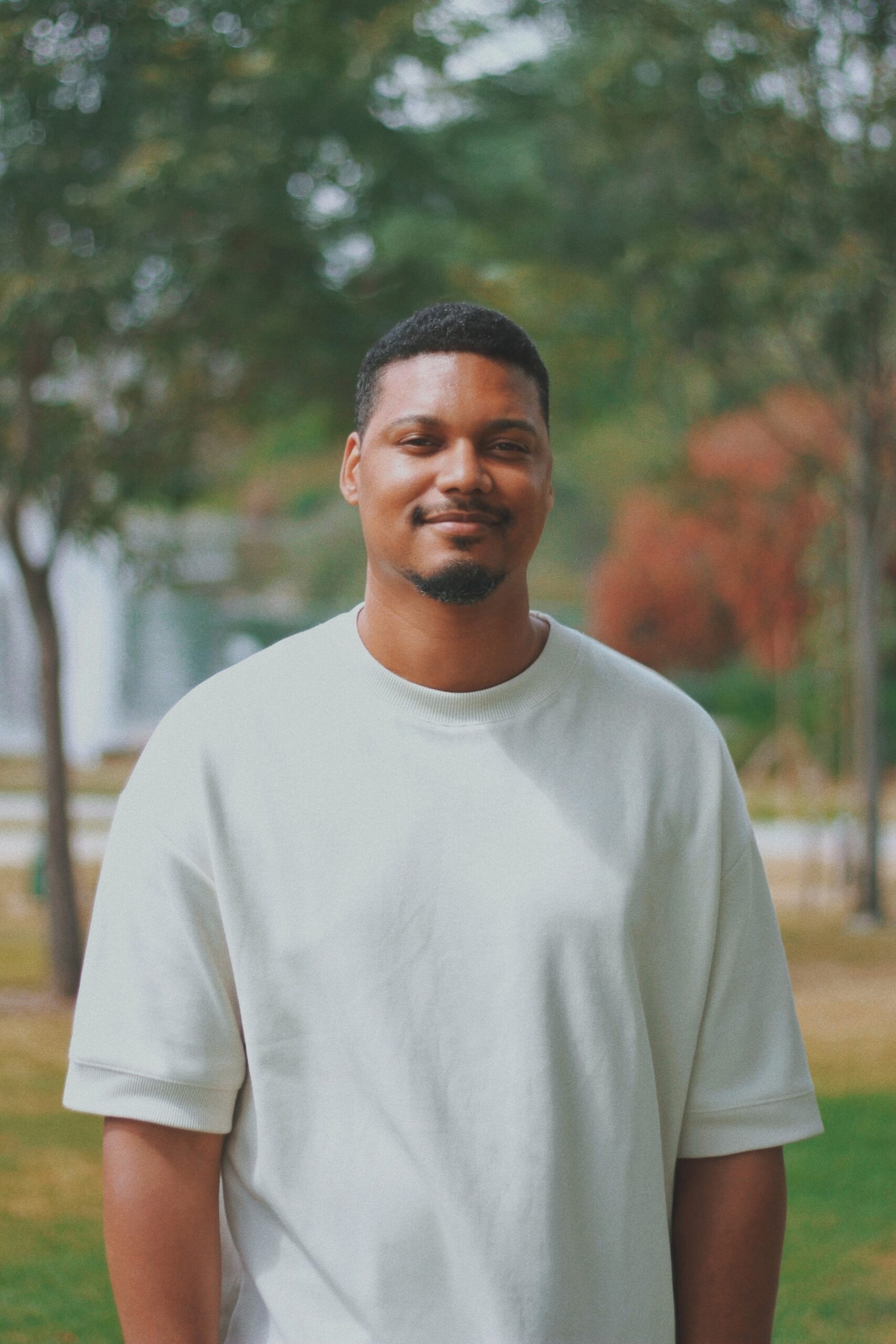 A man wearing a white t-shirt stands outdoors, smiling at the camera. Trees and greenery are in the background, suggesting a park setting. The lighting is natural and soft.