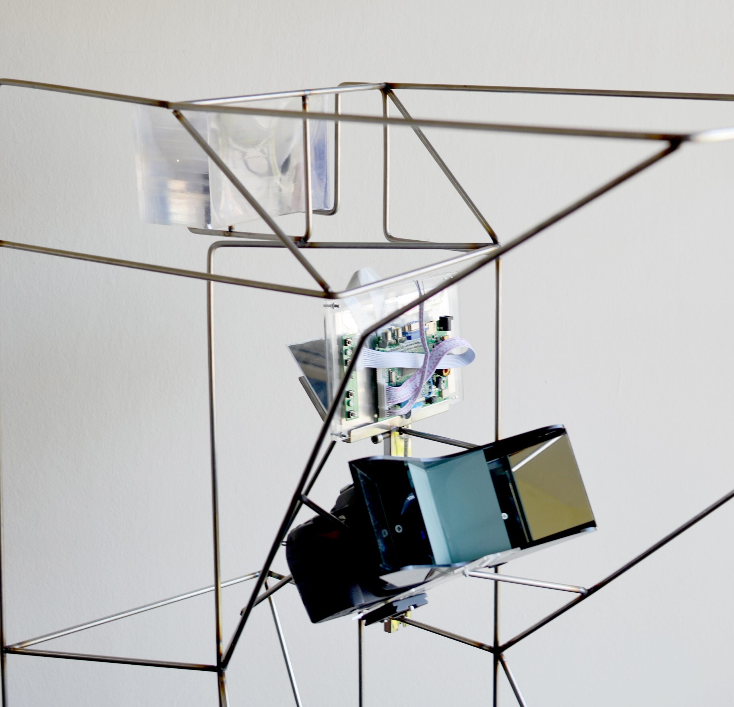 A geometric metal frame structure holds various electronic components, including circuit boards and angled reflective black panels, against a plain light background.