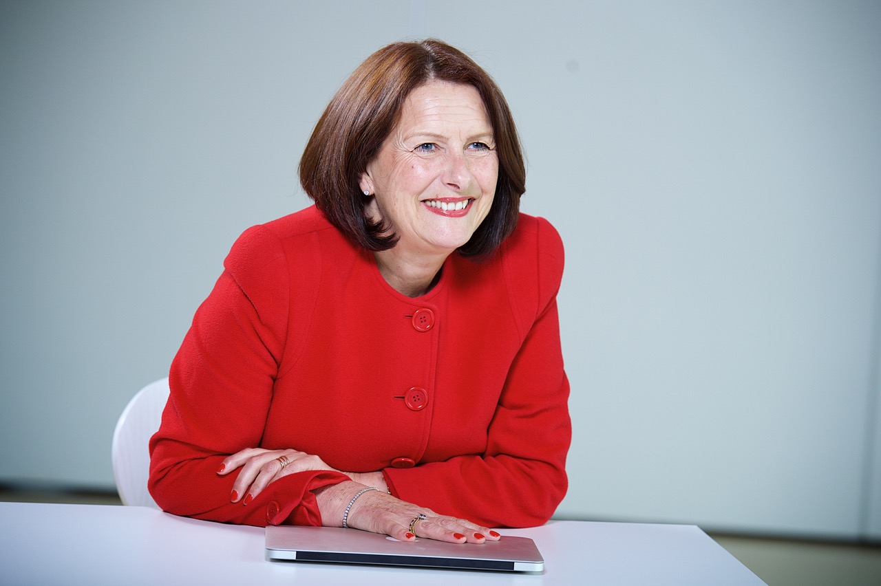 A woman with chin-length brown hair, wearing a bright red jacket, sits at a white table and smiles, resting her arms on a closed silver laptop. The background is plain and light-colored.