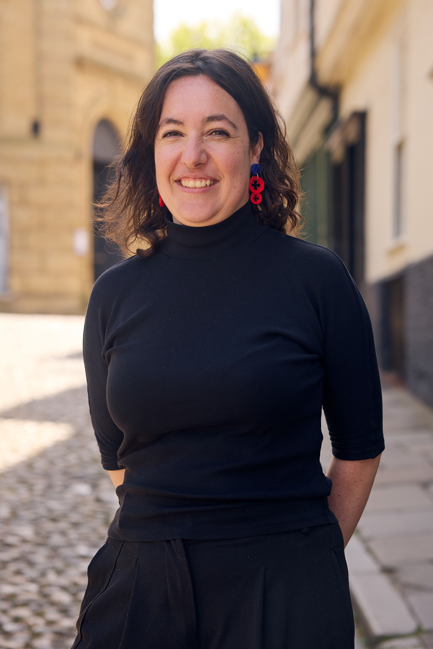 A woman with shoulder-length dark hair, wearing a black turtleneck and red earrings, smiles while standing on a sunlit cobblestone street with buildings in the background.
