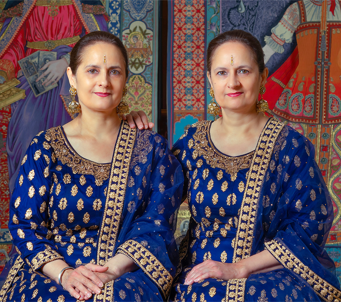 Two women dressed in matching royal blue and gold traditional outfits sit side by side with one arm around the other, posing in front of a colorful, ornate patterned backdrop.