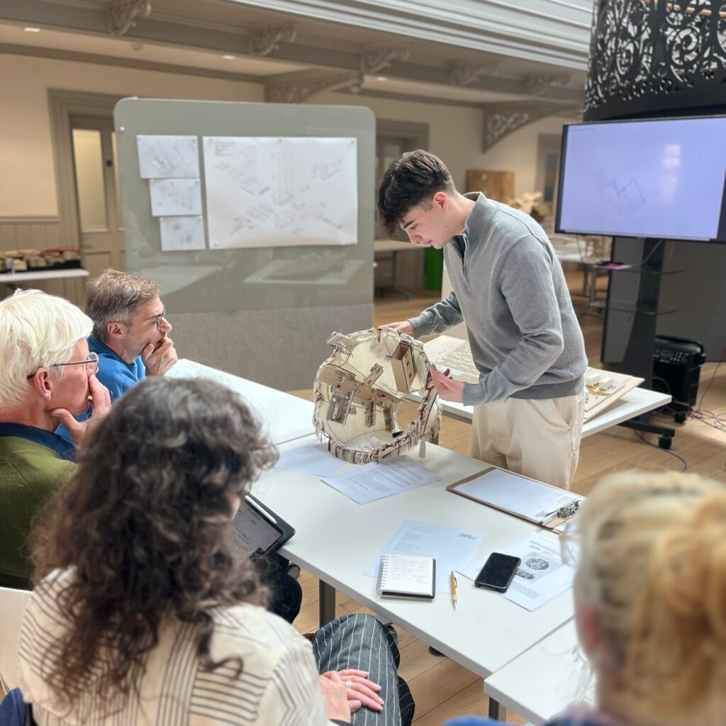 A young man presents a detailed architectural model to four seated judges in a modern room, with sketches and plans displayed on screens and boards behind them. Various notes and paperwork are spread on the table.